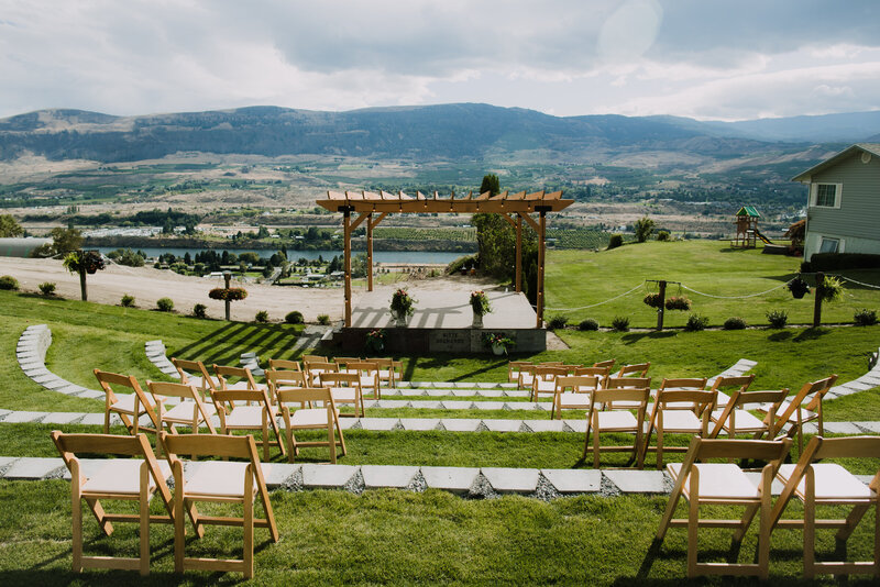 A beautiful view of the amphitheater at Union Hill Cider Co. in East Wenatchee, WA. Simple wooden folding chairs are set on each level of the amphitheater. There is a stage with flowers set on it. In the background there are beautiful foothills of the Cascade Mountains and the Columbia River.