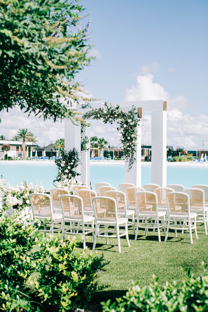 Rows of white cane-back chairs facing a white modern arch with greenery beside blue water at a Houston venue.