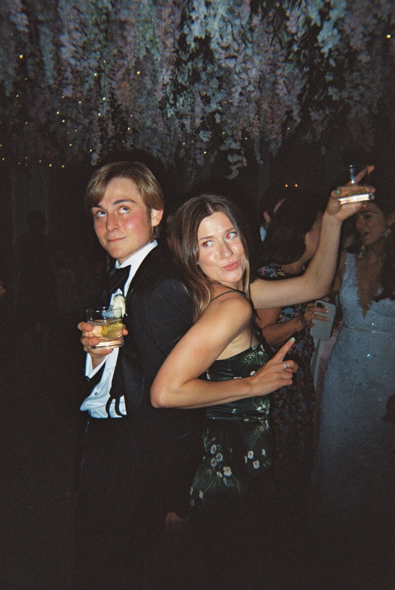 Flash photo of man and woman holding drinks dancing under a floral ceiling arrangement at wedding reception