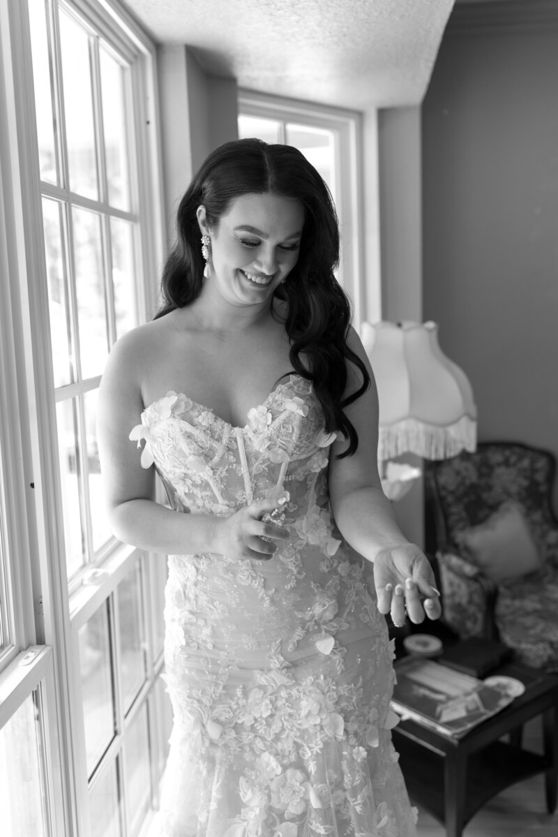 A bride in a lace gown standing beside a window as she gets ready, smiling softly in natural light—captured at an Oklahoma wedding planned by Harper Mae Events.