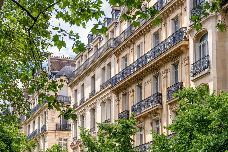 Elegant, historic European buildings with ornate balconies and large windows line a street. Lush green trees frame the foreground, with a partly cloudy sky visible overhead. The architecture is classic and sophisticated.