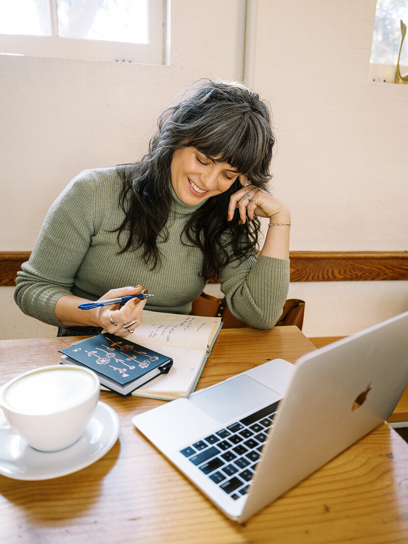 Bloem Creations founder Carrie smiles while writing in a notebook in a coffee shop, representing her love for copywriting services. 