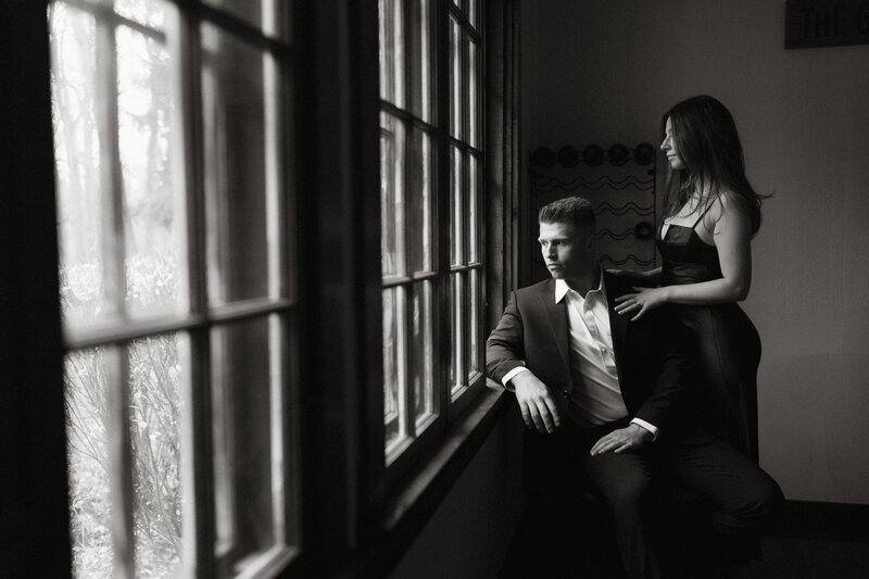 A couple posed by a large window in elegant black attire, sharing a quiet and cinematic moment together.