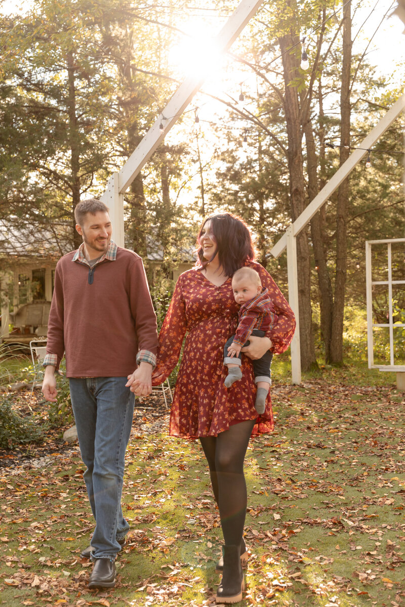 mom and dad hold hands and walk towards camera while smiling at each other, with mom holding baby son at Rustic Wild Arrow in the fall