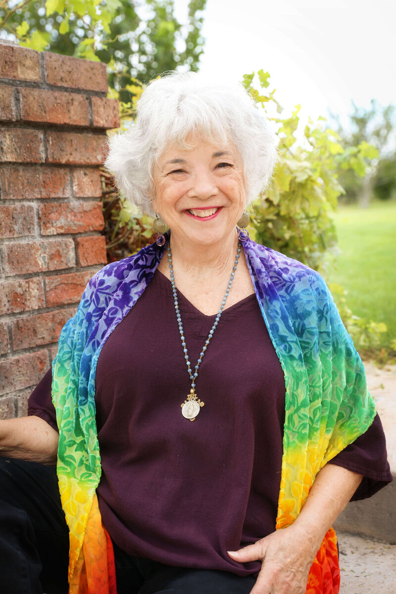 Dr. Samarah Grace wearing a purple blouse and rainbow shaw looking at the camera and smiling .  Green field behind her.