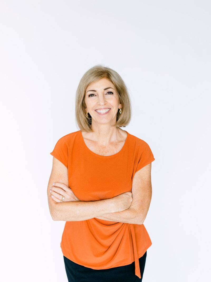Lora Ulrich smiling  wearing an orange top with crossed arms, radiating confidence.