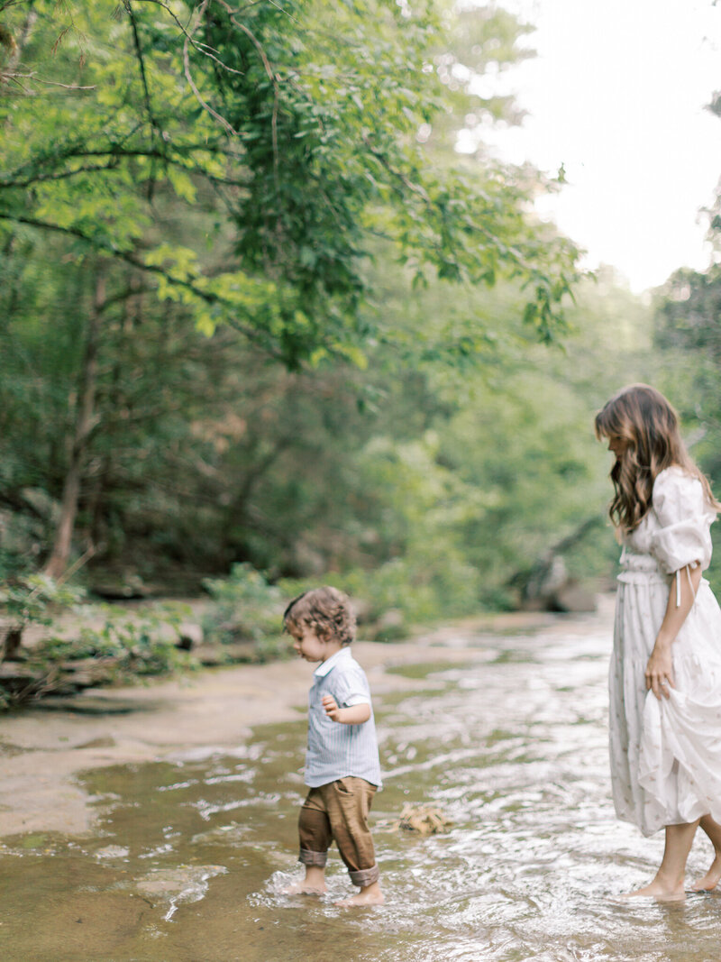 Brunette mother in white dress walks barefoot with curly-haired son in creek