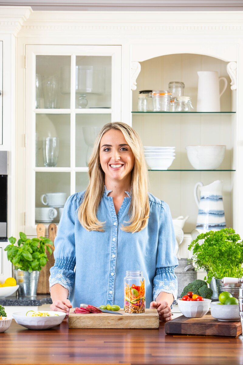 Charlotte Grand, Fertility and IVF Nutritionist, smiling in a bright, modern kitchen surrounded by fresh, colourful vegetables, herbs, and nutrient-rich ingredients. Representing The Fertility Kitchen’s focus on empowering women through fertility-friendly nutrition and lifestyle support