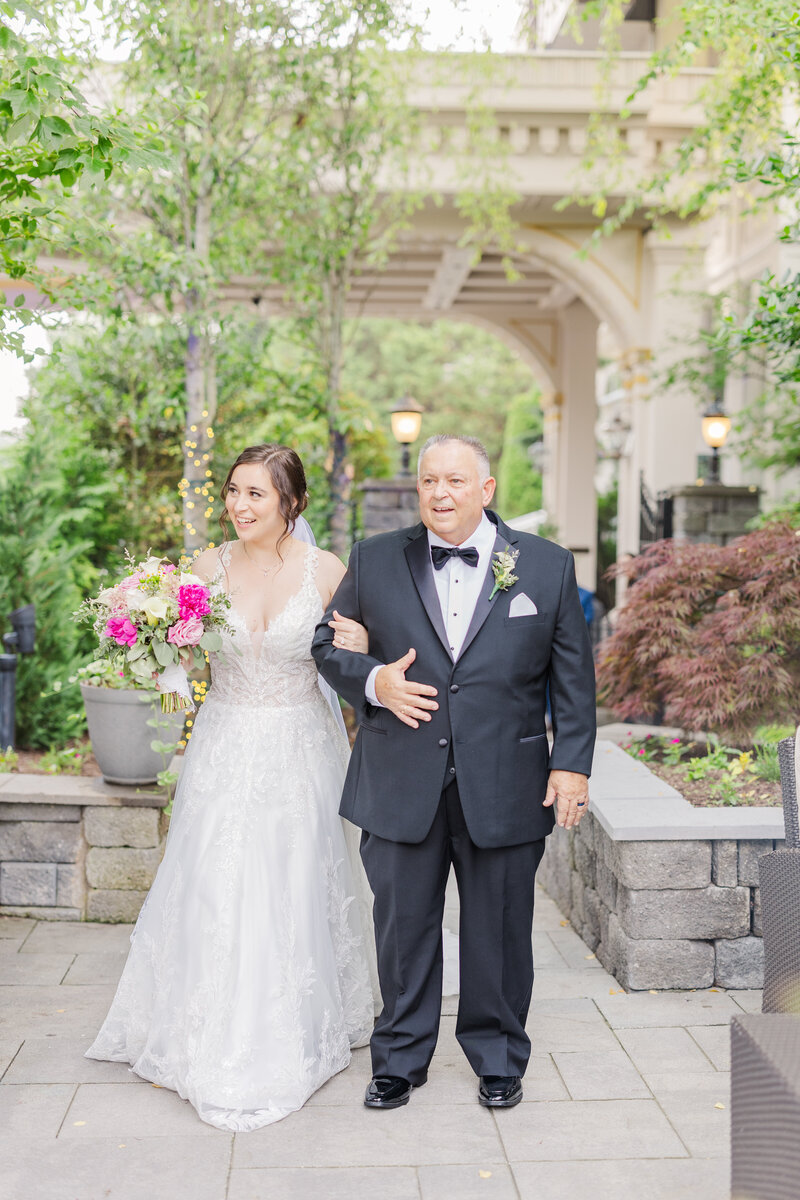 A father in a black tux walks his daughter down the aisle arm in arm for her wedding ceremony