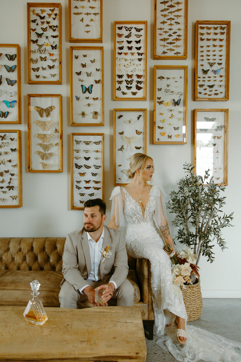 Bride and groom looking opposite directions sitting down on a leather sofa with butterflies behind them on the wall. 