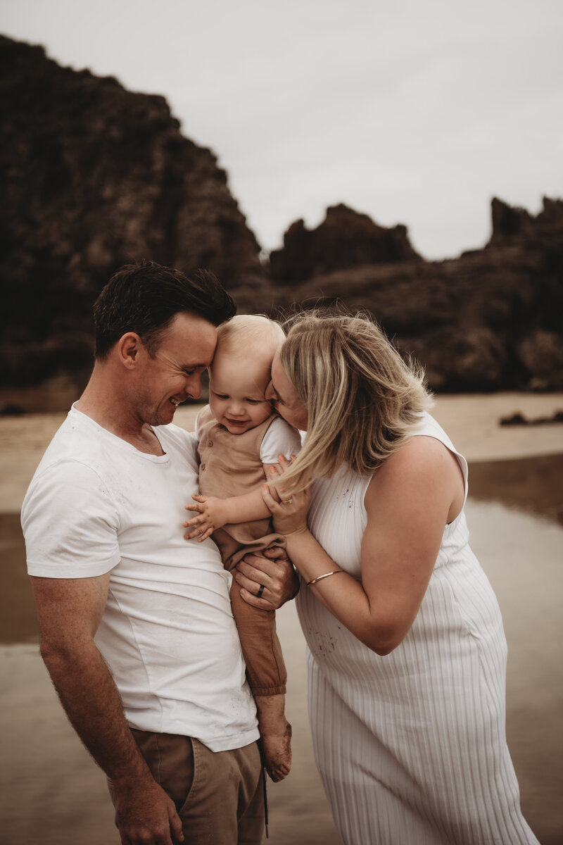 A man and woman stand on a beach holding a toddler between them. The adults are smiling, and the woman is kissing the child's cheek. Rocky cliffs are visible in the background, capturing the essence of Wollongong family photography perfectly.