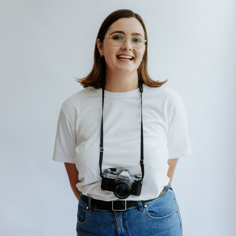 Anna Dewar smiling to the camera wearing a Canon ae-1 around her neck.