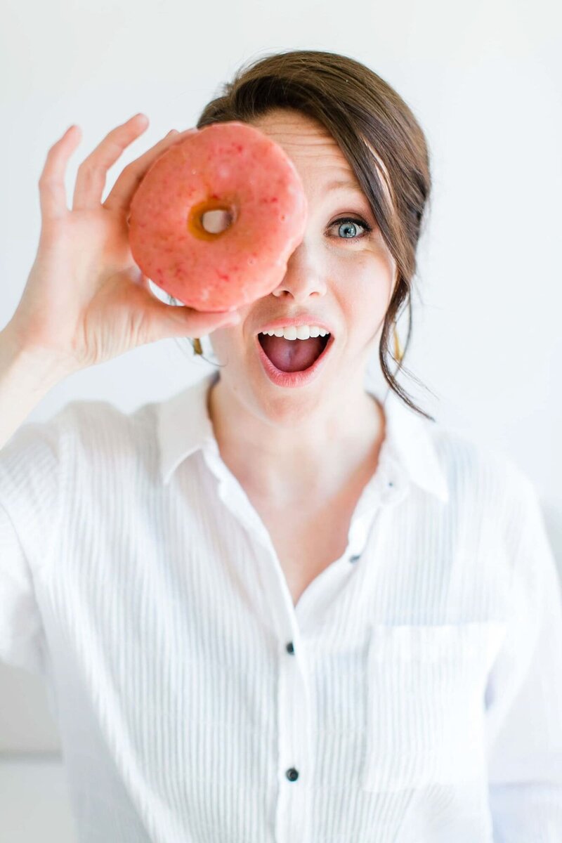 honeybook pro caitlyn gray holding a pink donut over one eye and smiling because she is offering a honeybook coupon code to users