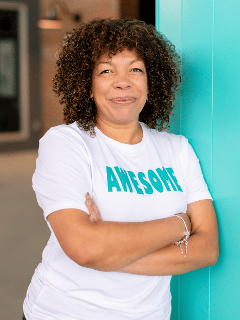 Photo of Michelle McKown -Campbell smiling while wearing a white t-shirt with AWESOME  in teal writing while leaning against a teal wall with her arms crossed