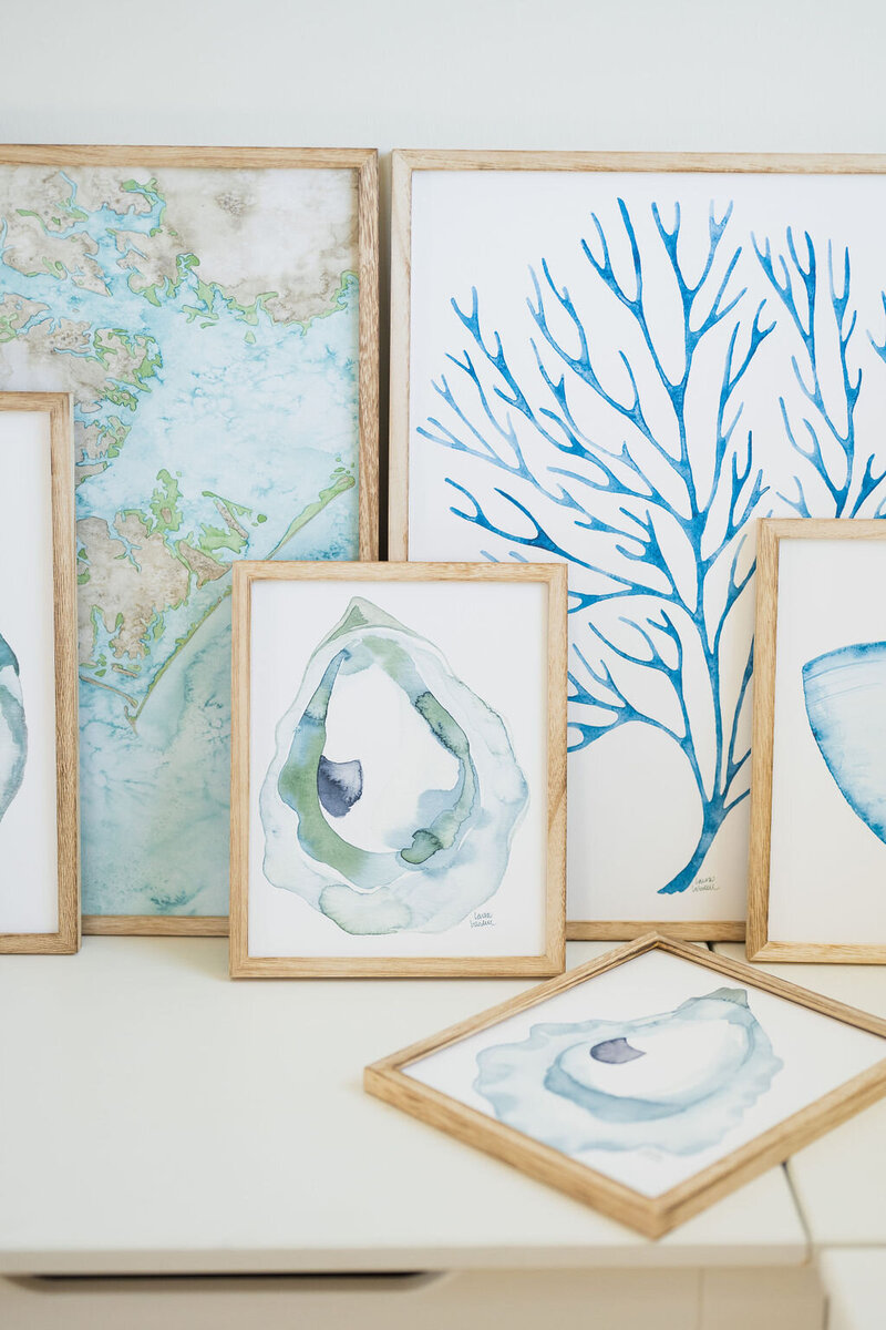 coastal watercolor artiwork in hues of blue with wooden frames leaning against a white wall in wilmington, north carolina