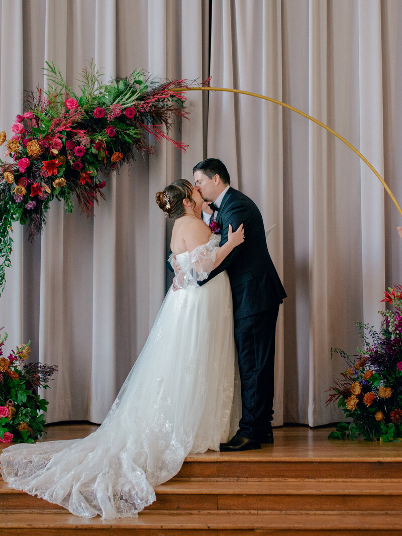 A bride and groom kiss under a floral arch after their wedding ceremony.