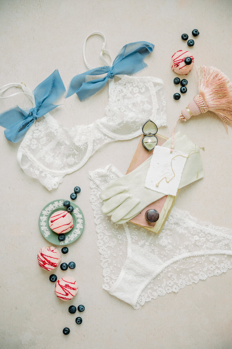 Richmond Boudoir Photography | White floral lace lingerie with blue ribbon tied on the straps laying amidst little cakes, blueberries, a vintage book, and a pair of cream vintage gloves.