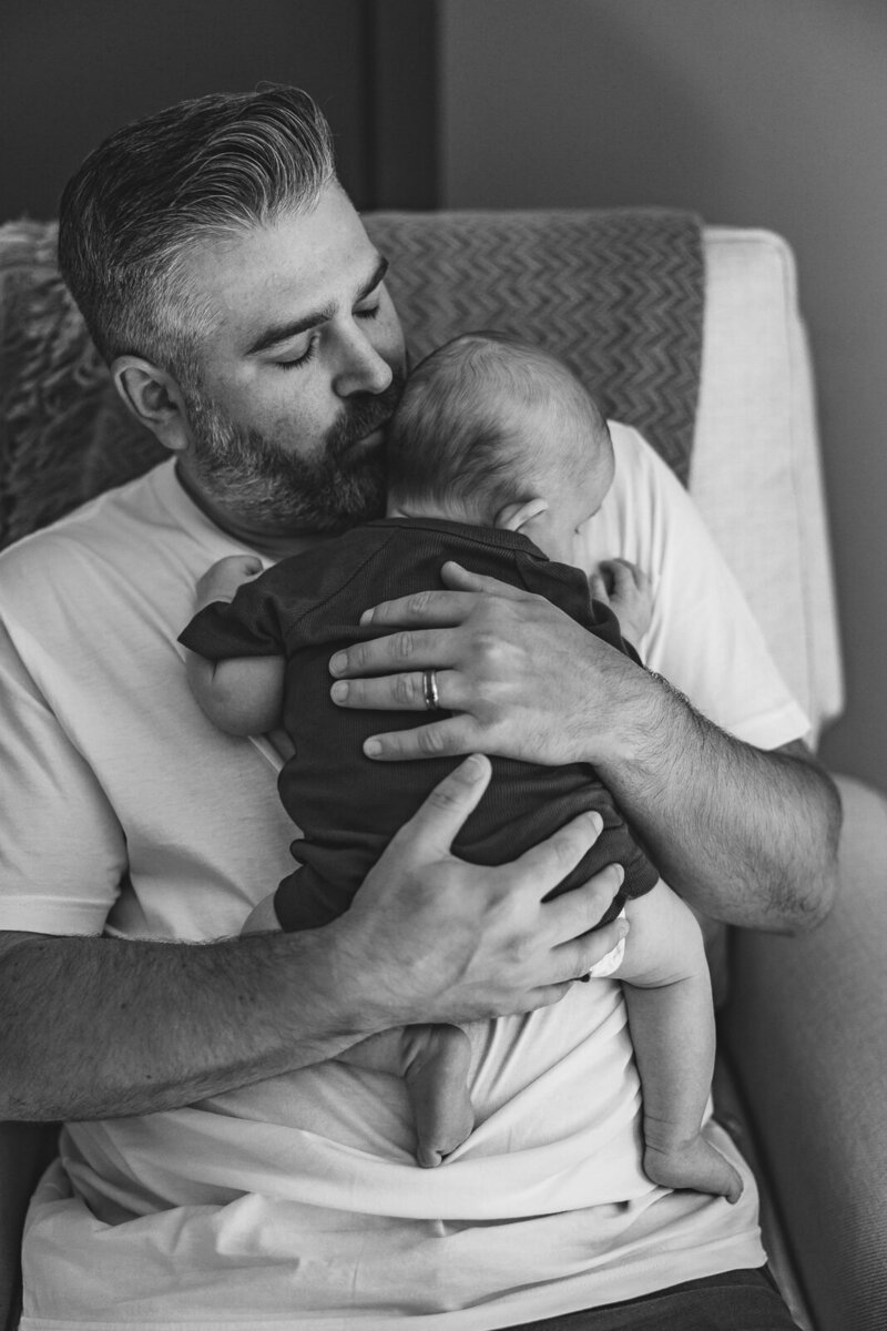 A black and white portrait of a dad kissing his infant son's head while sitting in a chair during a newborn session in St. Louis, Missouri. 