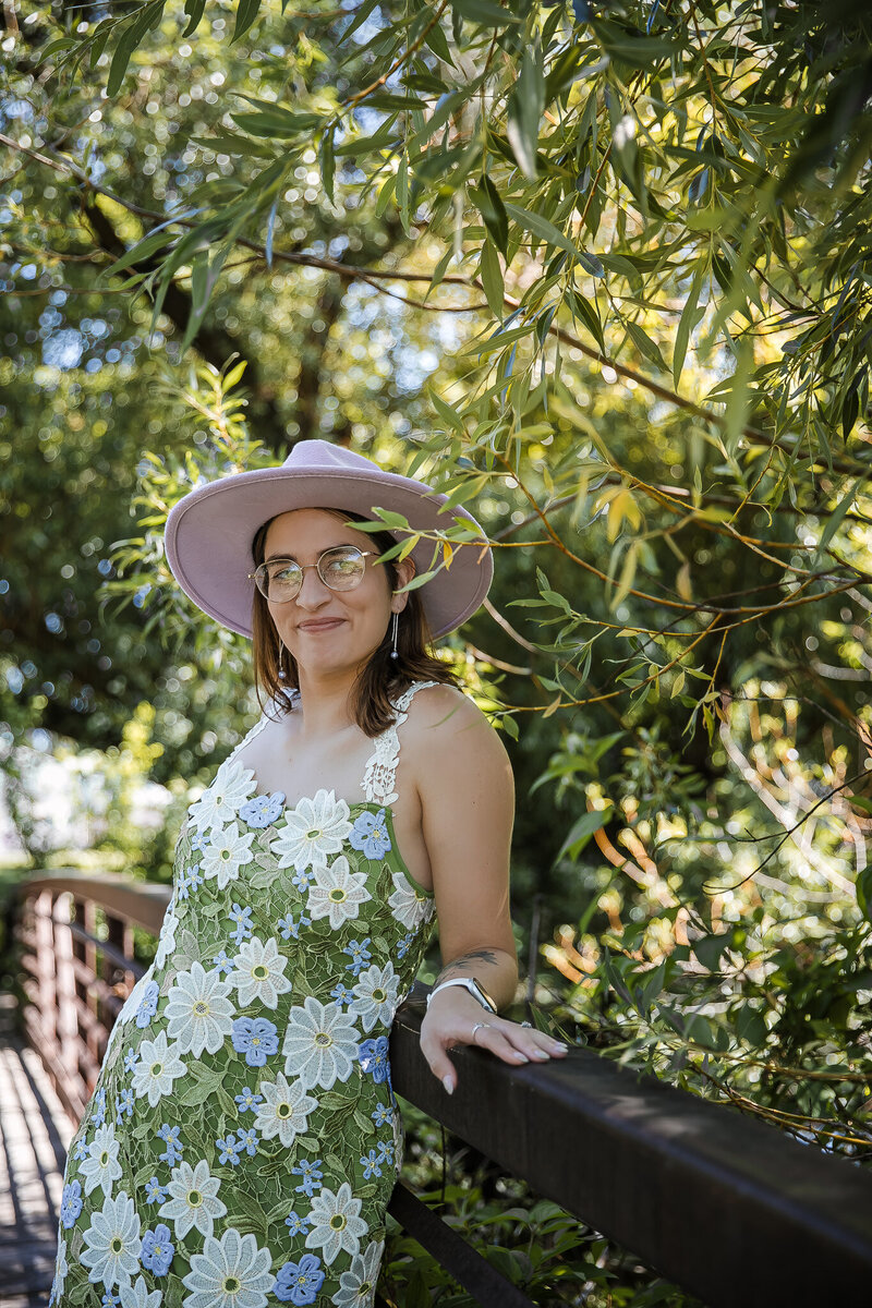 A woman with brown hair and glasses wears a purple felt hat and a floral embroidered dress posing on a bridge with willow branches