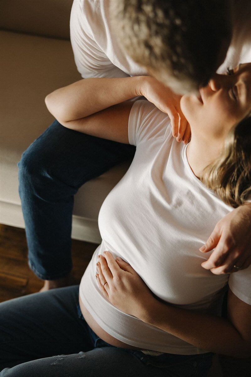 Woman in white tee and light wash denim jeans holds her pregnant belly as husband goes in for a kiss