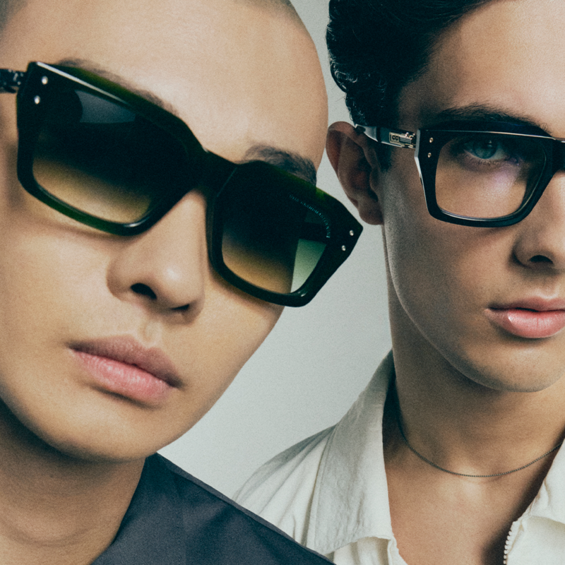 Two men, each wearing luxury eyewear. One man has sunglasses and the other has bold standard glasses.