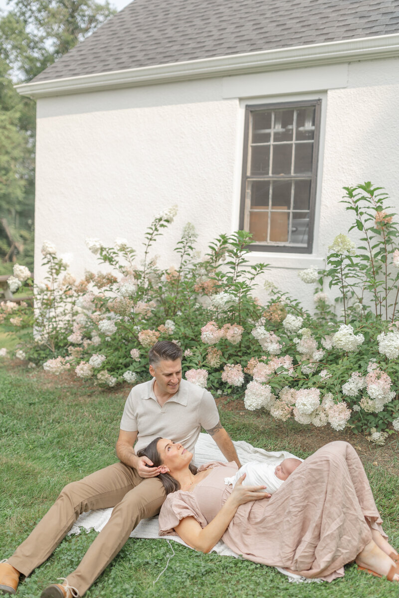 A tender outdoor newborn session featuring a mother and father surrounded by beautiful flowers. The husband sits on a blanket, gently cradling his wife's head in his lap as she gazes lovingly at their newborn resting in her arms. The soft natural light and serene setting create a warm, intimate family moment