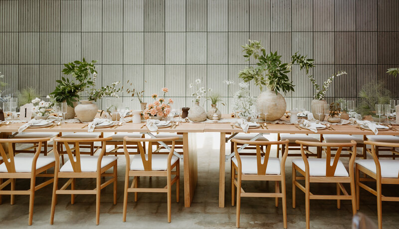 Long wooden banquet tables styled with natural wishbone chairs, neutral pottery, and greenery in a contemporary Houston venue.