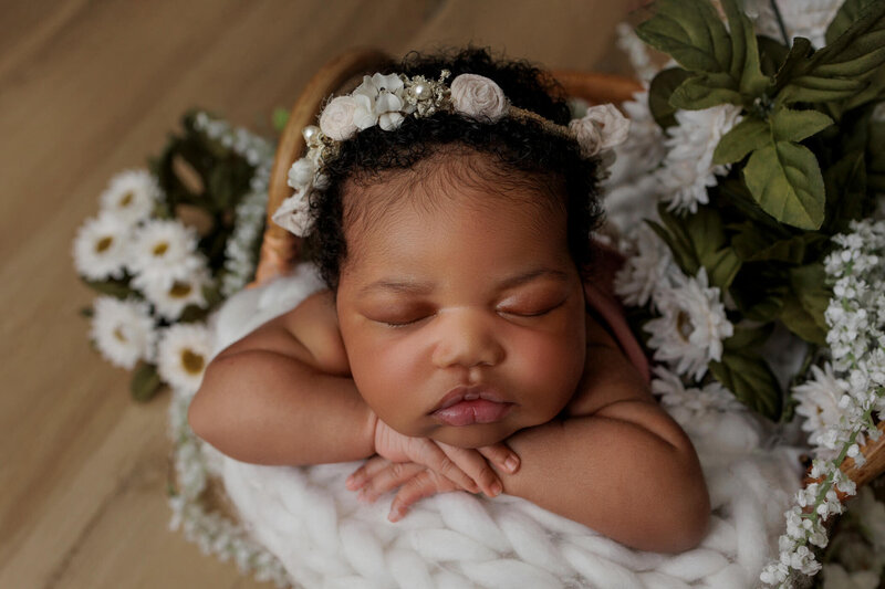 A close-up of a newborn baby girl with a floral headband, sleeping peacefully on a soft, white knitted blanket with flowers in the background.