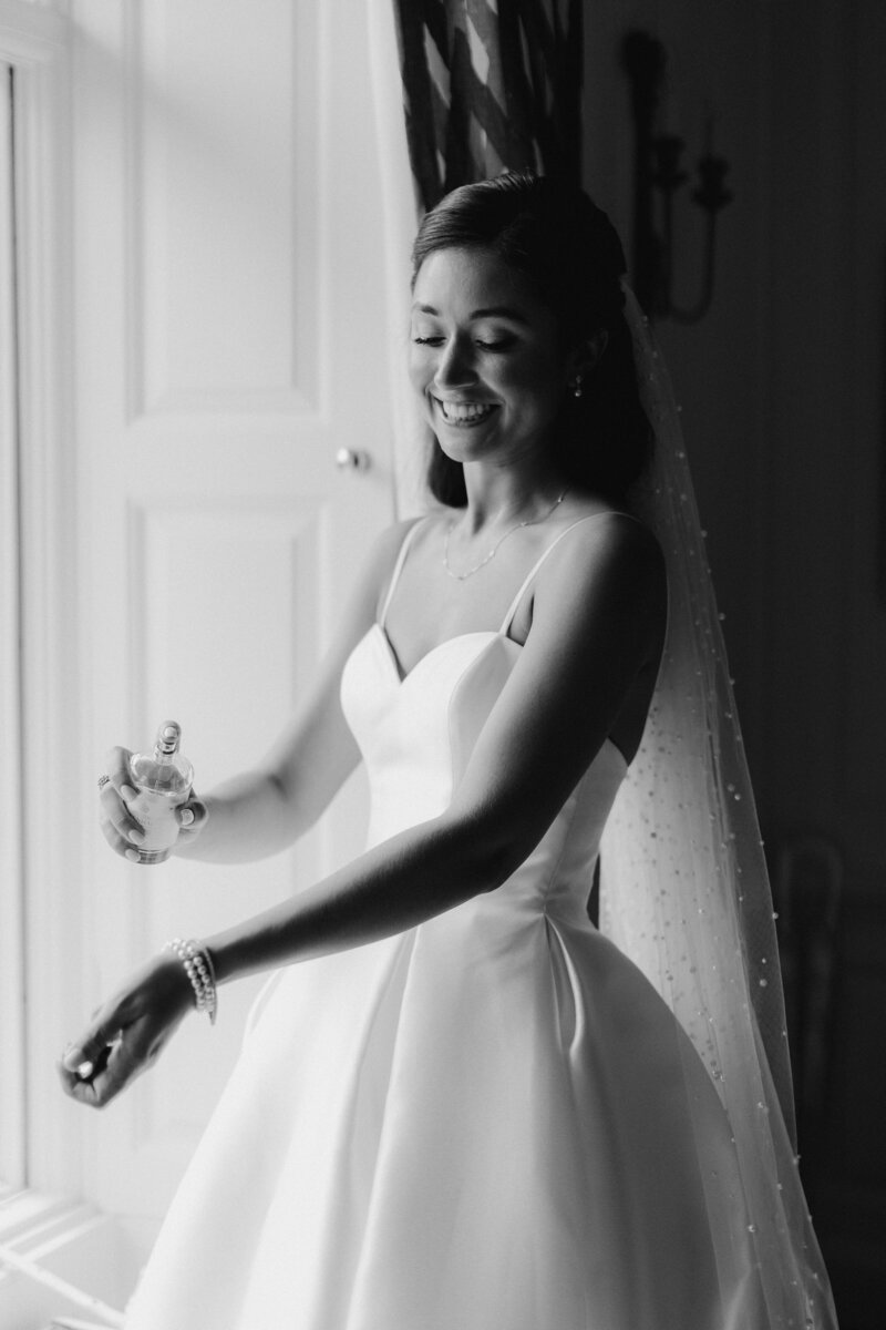 Soft black and white wedding photo of bride during bridal prep spraying her Penhaligons Empressa perfume by the window light