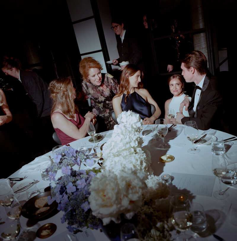 A joyful and intimate moment captured at the bride and groom's wedding reception at Cutler and Co. The bride, seated at a round table, is wearing a sleek white halter-neck gown and smiling warmly as she interacts with guests. The groom, dressed in a classic black tuxedo, leans in affectionately towards the bride, holding her close as they enjoy the evening together. Around the table, friends and family in elegant attire share in the celebration, laughing and talking with one another. The table is adorned with luxurious floral centerpieces of white hydrangeas and soft purple blooms, with wine glasses and plates scattered across, reflecting the casual yet refined nature of the evening. The ambient lighting casts soft shadows, creating a warm and inviting atmosphere for this chic wedding celebration at a high-end Melbourne venue.