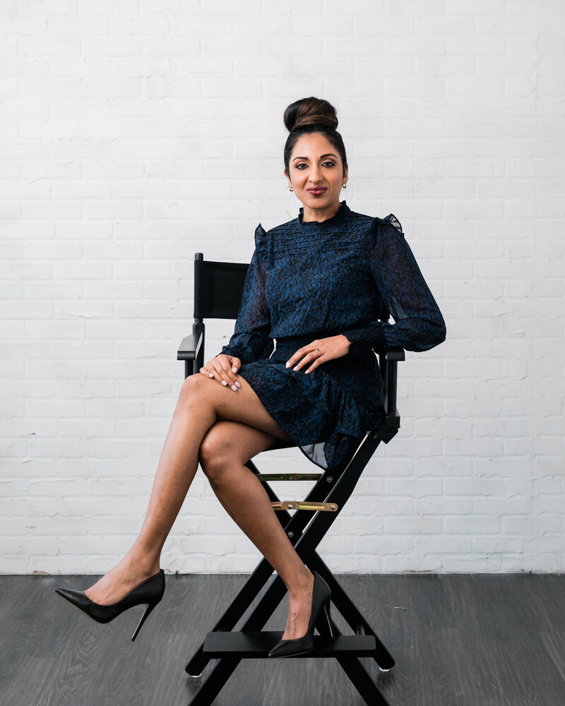 Priya Sam, Toronto Keynote Speaker and Podcast Host, poses in director's chair wearing a navy blue dress.
