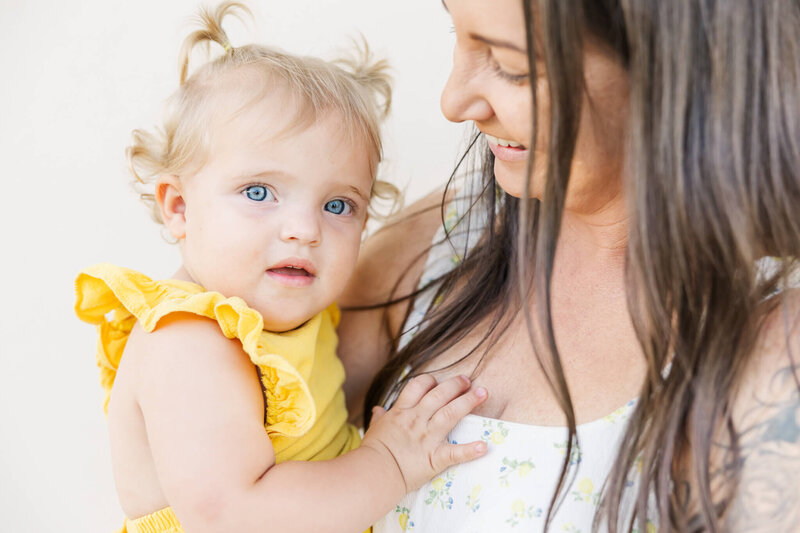Toddler girl wearing a daffodil yellow with beautiful blue eyes is being held by her brown haired mum who is wearing a floral dress with yellow flowers.