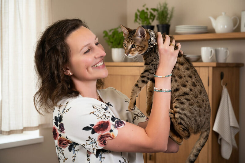 A smiling woman in a floral shirt holds up a spotted cat in a cozy kitchen with wooden cabinets, white dishes, and potted plants—reflecting the warmth found with a therapist for teens and adults in Orange County.