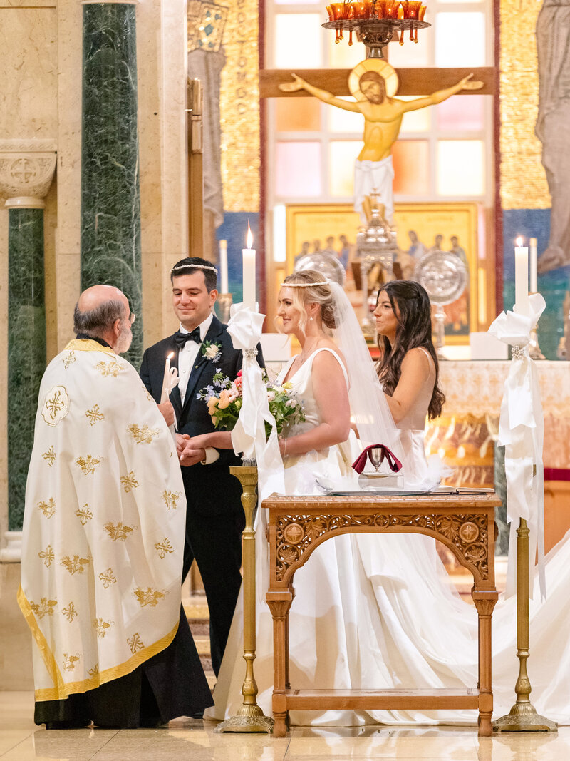 A bride and groom stand hand in hand in a church during their wedding ceremony. The priest, in traditional robes, leads the service. A bridesmaid stands beside the bride, holding a bouquet. The church interior is ornate, with a crucifix in the background.