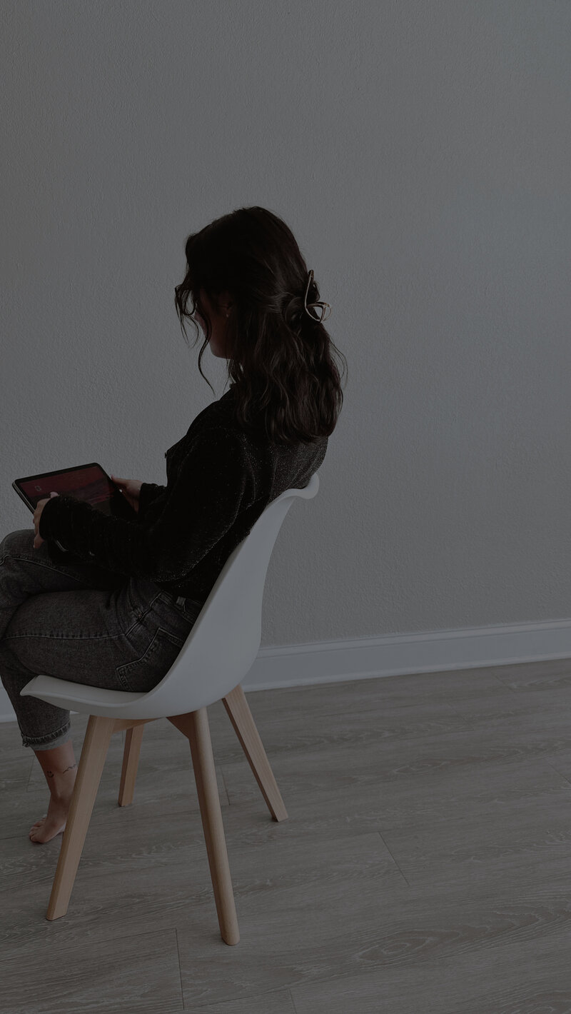 A woman with wavy hair sits on a white chair in a minimalist room, focused on a tablet. The setting is calm, with neutral tones and wooden floors.