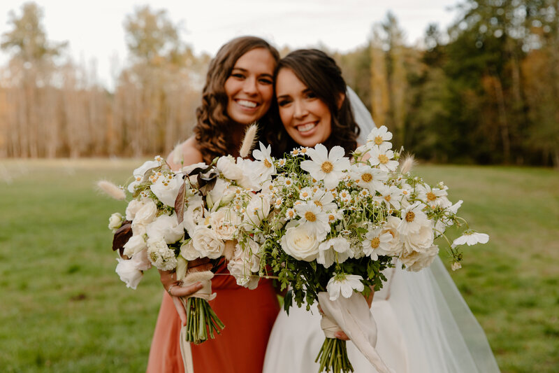 Bride and maid of honor hold up bouquets to the camera in a field