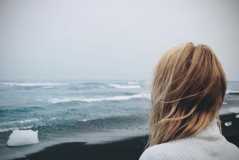 Langhaarige Frau in kuscheliger Strickjacke am Strand, das Gesicht vom Wind verdeckt, während die Haare in alle Richtungen wehen - ein Moment der  Selbstreflexion und des Aufbruchs