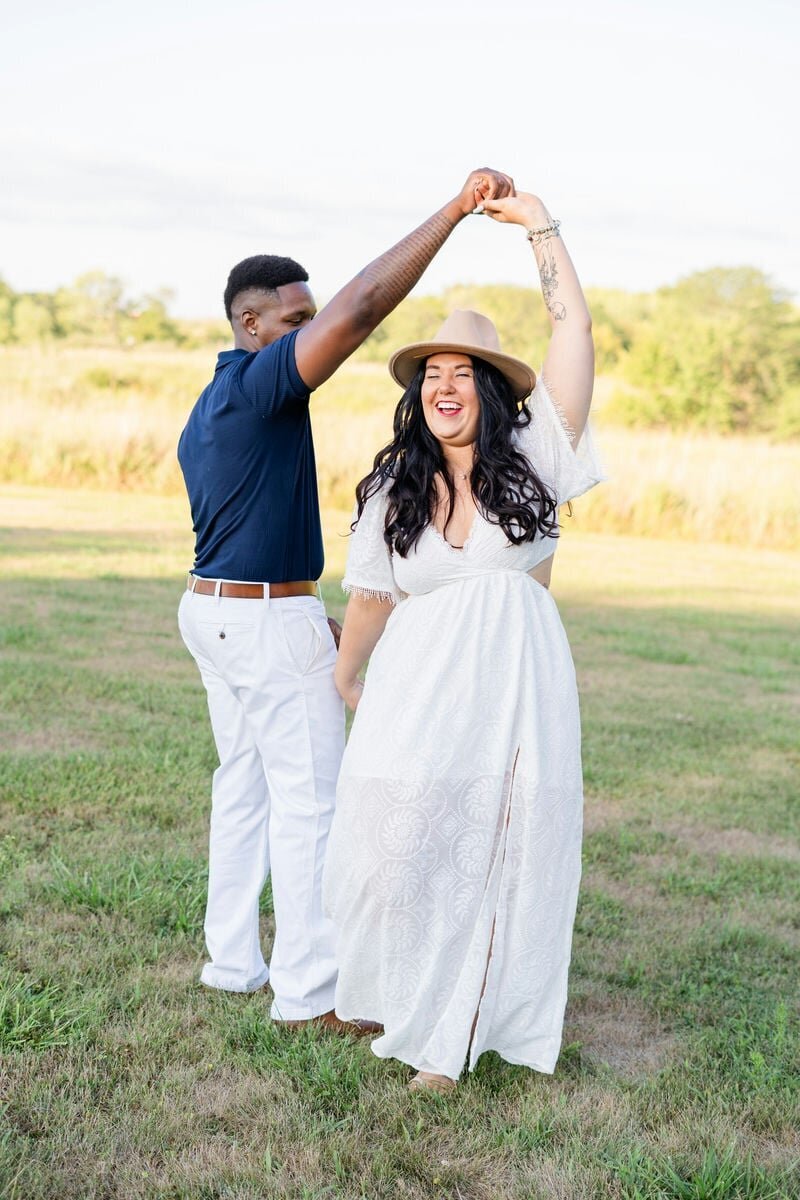 A groom twirling his future wife in the middle of a field, smiling.
