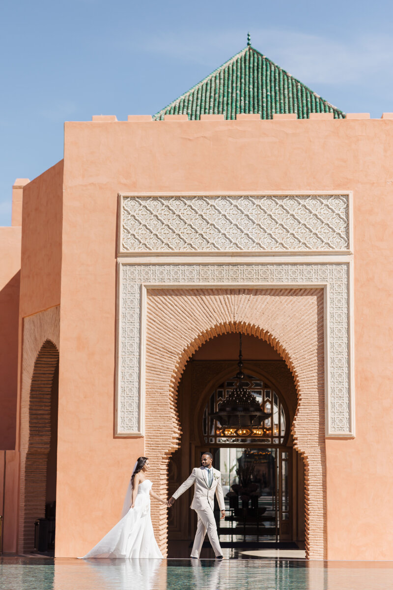 bride and groom walking in front of peach buiding at the oberoi in Marrakech