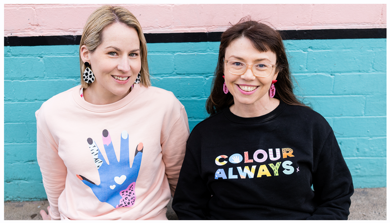Crystal Oliver and Jessica Roberts looking cool and smiling at the camera wearing a pink jumper with a hand and a black jumper with colourful letters saying colour always