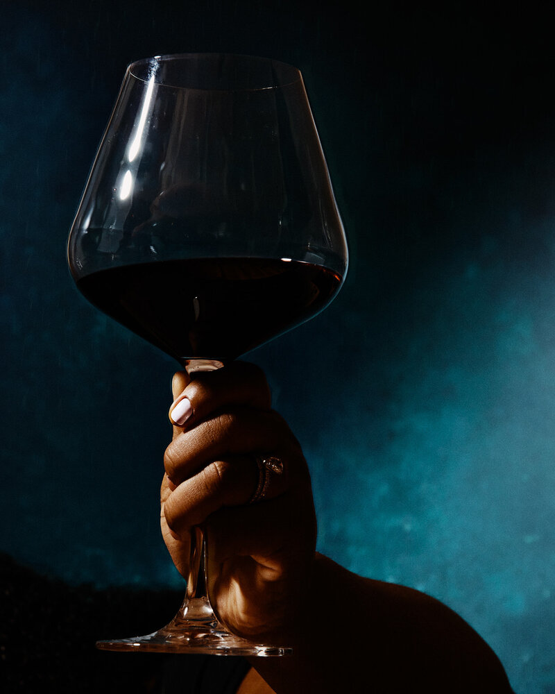 A powerful and dramatic image of a hand holding a glass of red wine against a deep blue background. Captured by Mica McCook, this photograph emphasizes the richness and boldness of the wine, creating a striking visual impact.