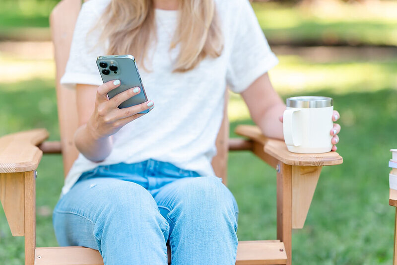 Person uses an iPhone and drinks coffee while sitting on a chair in the park