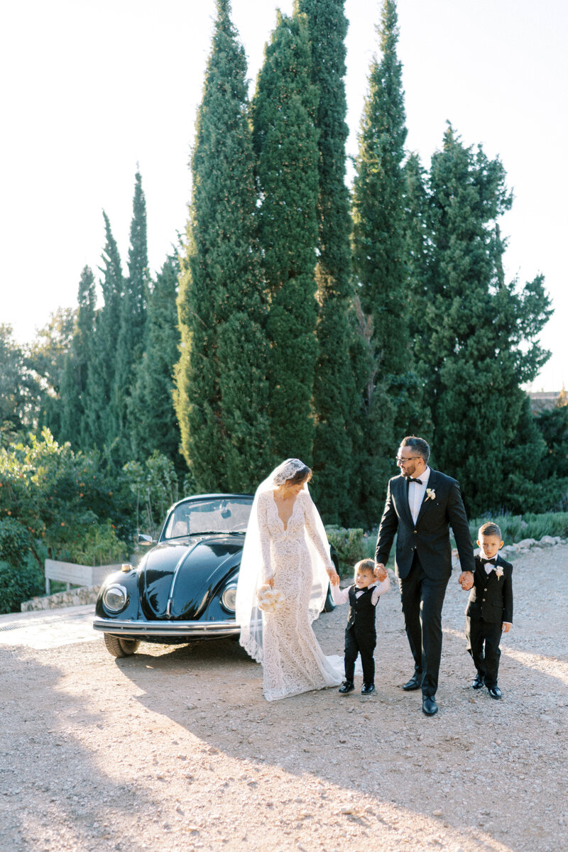Bride and groom walking hand in hand with their children in formalwear beside a classic black car on their wedding day