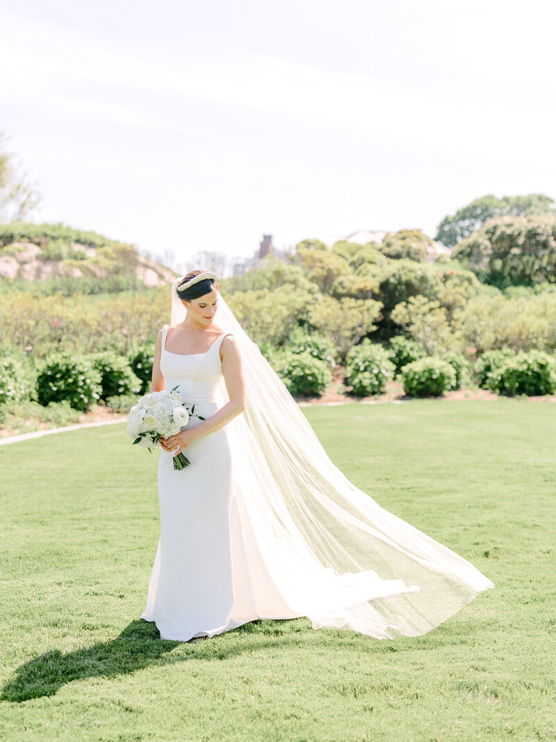 Bride standing in the grass holding her bouquet with her veil blowing behind her