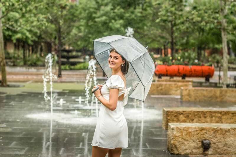 High School graduate in white dress holding umbrella in rain walking through The Pearl downtown San Antonio