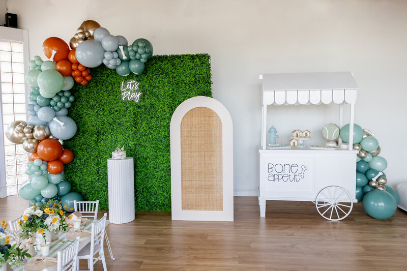 Party setup with floral wall, rattan arch, dessert cart and cake stand.