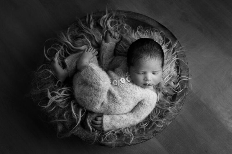 Newborn baby in a knitted onesie  laying on light coloured fur in a wooden bowl.