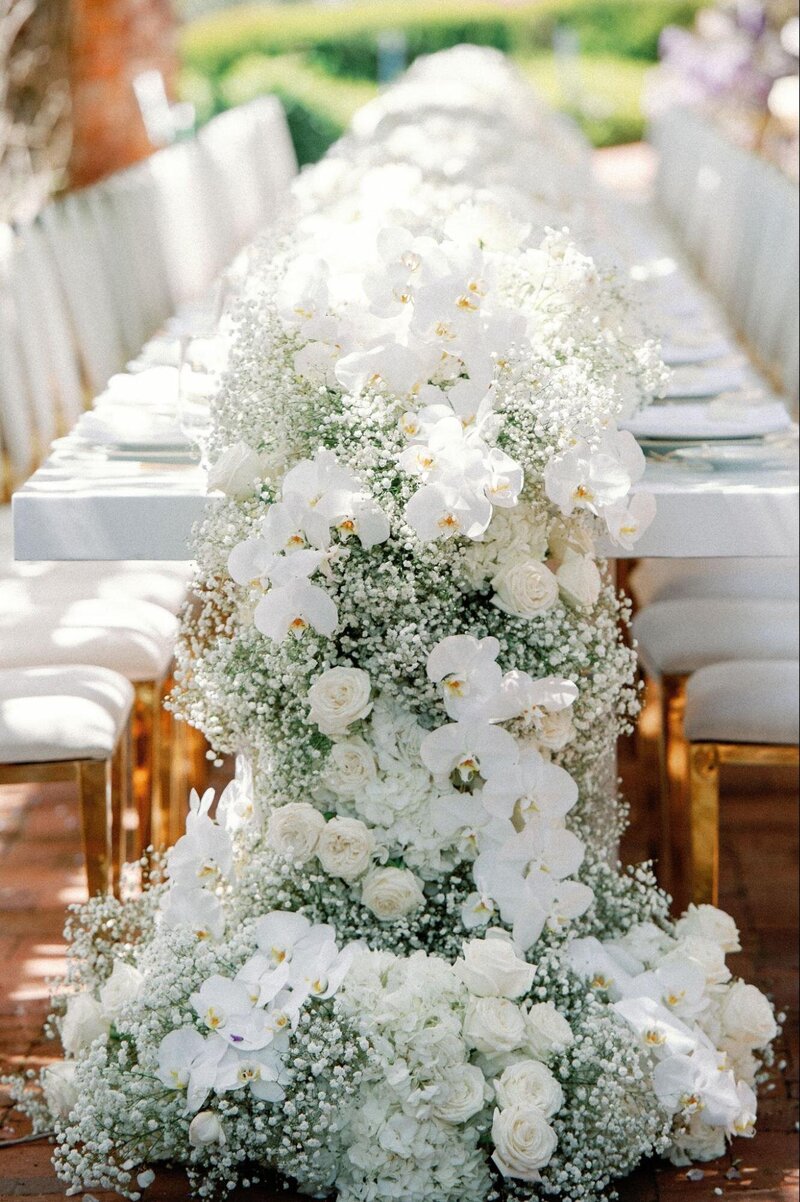 A spray of white flowers gracefully extends beyond the length of a white reception table and onto the ground