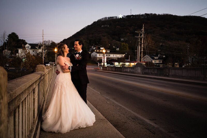 A groom stands behind a bride wearing a wedding dress and both of them smile as they get into a pose set by their Virginia elopement photographer. They're photographed on a bridge in Roanoke, Virginia with Mill Mountain and the Roanoke Star illuminated behind them.