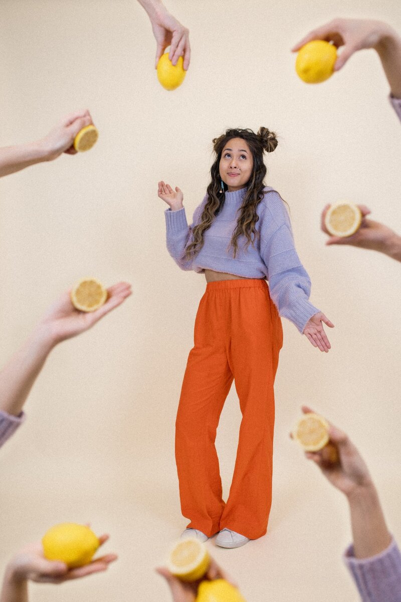 Woman with half-up pigtail buns wearing a purple top and orange pants poses. Several other hands frame the photo holding half and whole lemons.